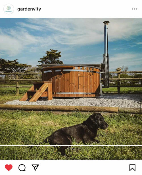 Wood-fired wooden hot tub installed on gravel with a scenic rural fence backdrop and a black dog resting on the grass in foreground.