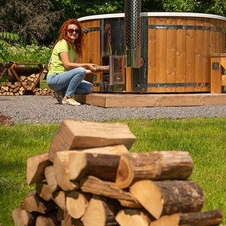 Outdoor view of woman lighting fire in wood fired hot tub stove surrounded by firewood and greenery background