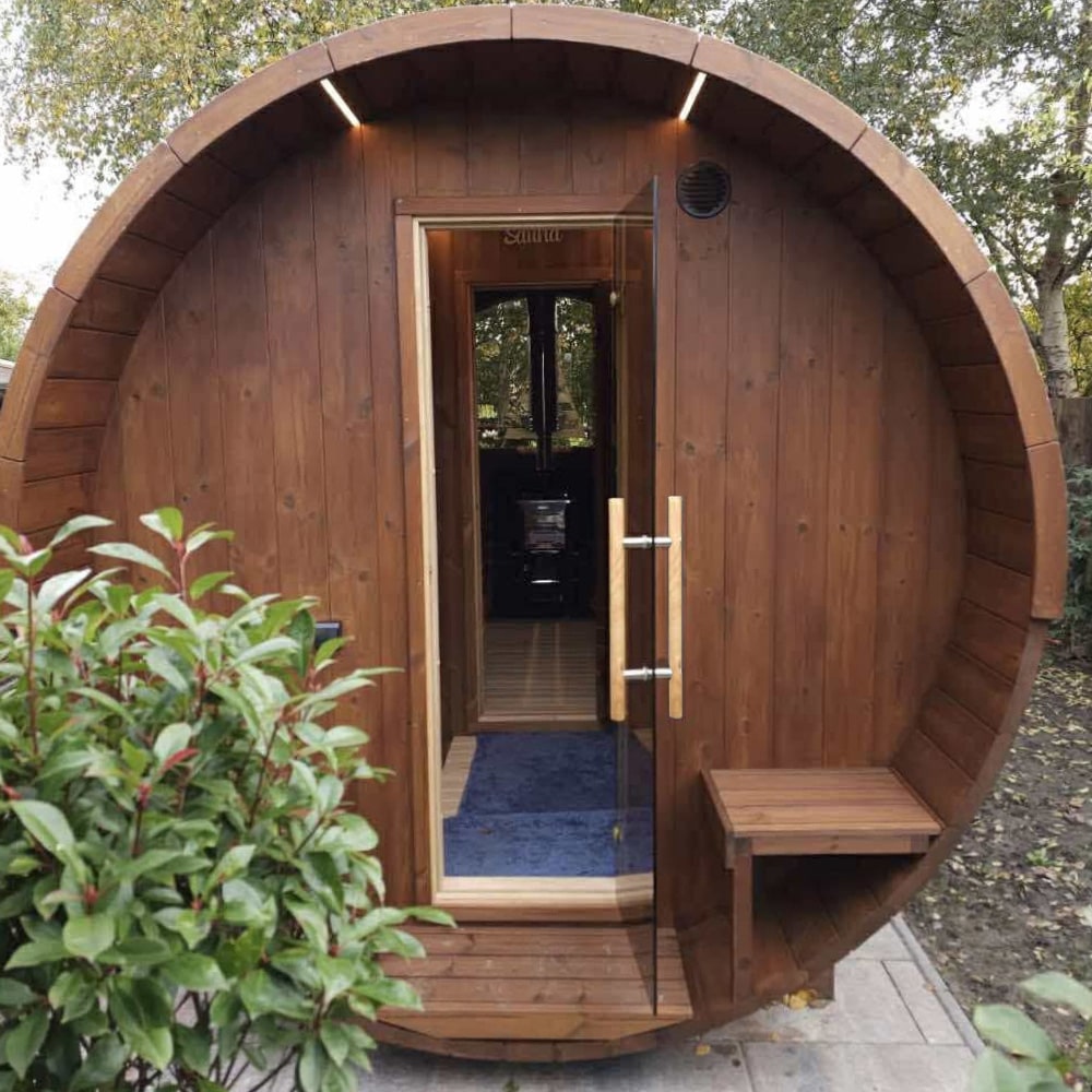 Front view of a barrel-shaped wooden sauna with glass door, small exterior bench, and surrounding garden foliage