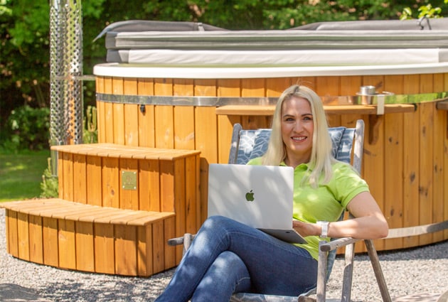Woman sitting on a chair using a laptop in front of a wood-fired hot tub with wooden steps outdoors