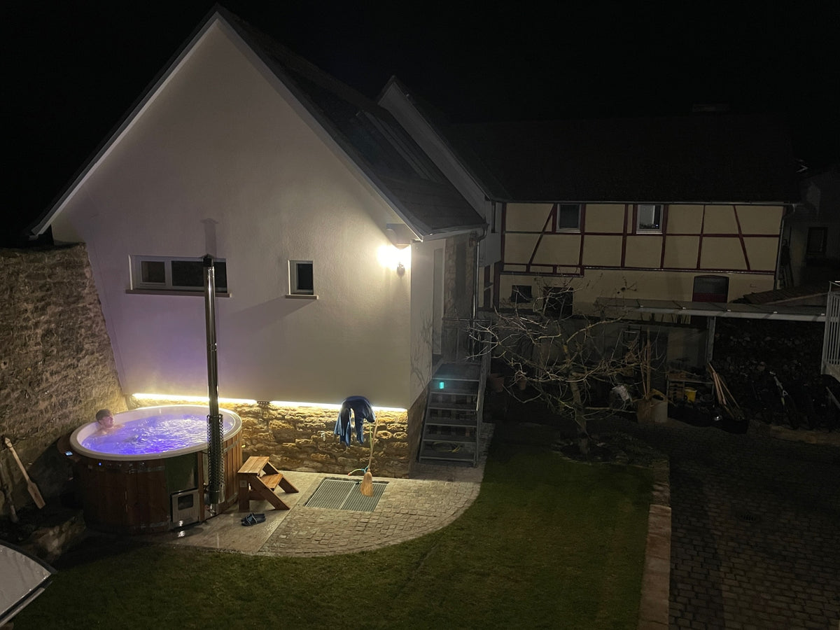 Nachtansicht eines beleuchteten Außen-Whirlpools neben einem Stein- und Putzhaus mit angrenzendem Garten und Holztreppe