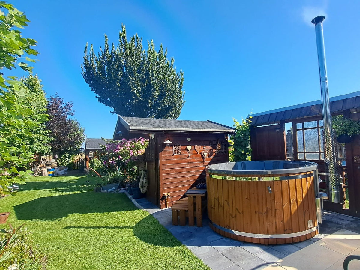Außenansicht eines runden hölzernen Whirlpools mit Kamin neben einem Holzschuppen und einem grünen Garten unter klarem, blauem Himmel