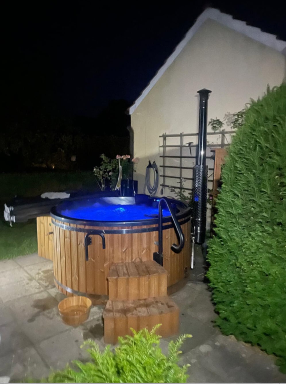Nachtansicht eines hölzernen Whirlpools mit blau beleuchtetem Wasser, Holzstufen, Edelstahl-Ofenrohr und Gartenumgebung