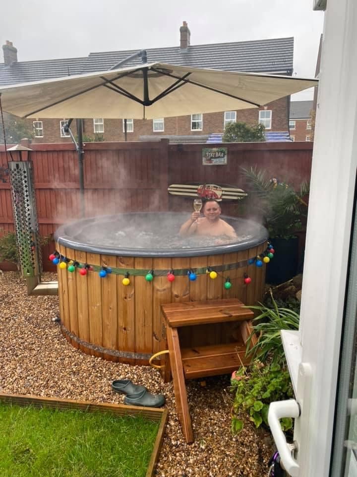 Außenansicht eines runden, holzbefeuerten Hot Tubx mit Holzstufen, bunten Lichtern, großem Sonnenschirm und aufsteigendem Dampf in einer Hinterhofkulisse