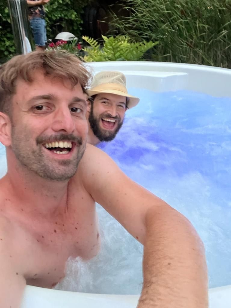 Zwei Männer lächeln und entspannen sich in einem sprudelnden Whirlpool im Freien, umgeben von Grün, bei Tageslicht.