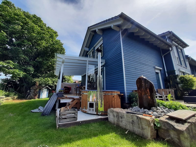 Seitenansicht eines blauen Holzhauses mit überdachter Terrasse, Außenheizung, Brennholzstapel und umliegendem Garten mit grünem Rasen