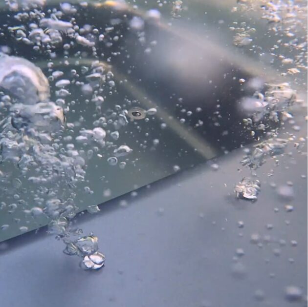Nahaufnahme unter Wasser von aufsteigenden Luftblasen vor einem verschwommenen grauen Oberflächenhintergrund.