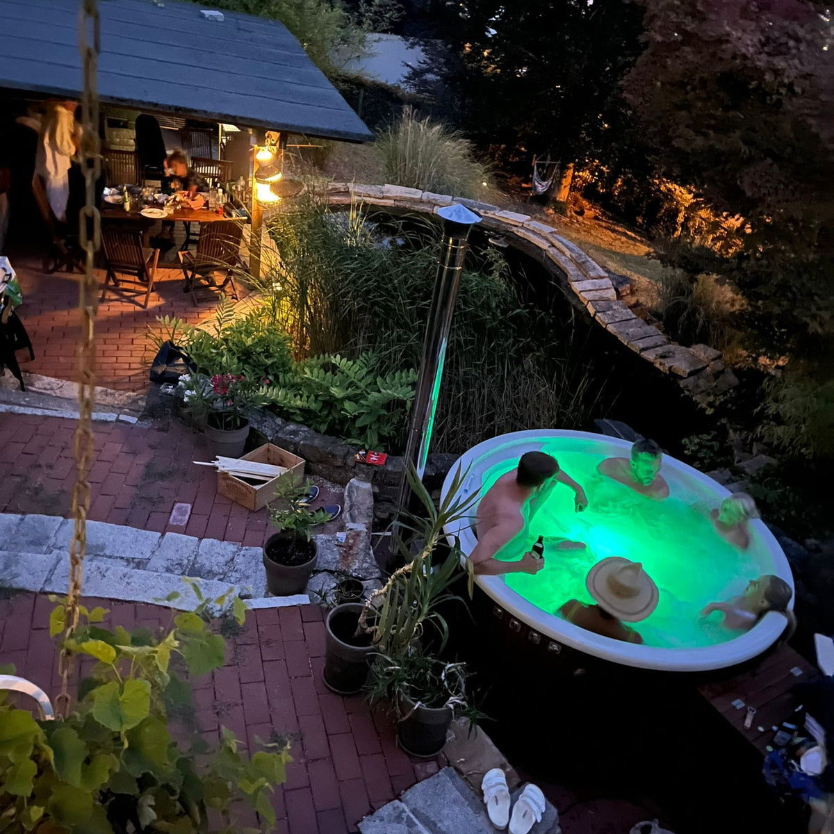 Abendlicher Blick auf einen holzbefeuerten Whirlpool, der grün leuchtet, während vier Personen auf einer Gartenterrasse mit Pflanzen und Sitzgelegenheiten entspannen.